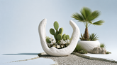 A white crescent-shaped planter holding a green prickly pear cactus and small white flowers, styled on a light stone surface beside a larger white planter with a palm tree and succulents against a pale blue background.